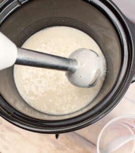 an immersion blender being held over a white liquid in a slow cooker